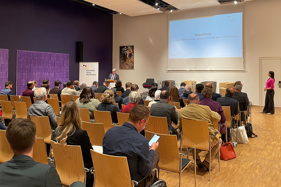 Ein Männ steht hinter einem Rednerpult vor einem Publikum auf Stuhlreihen. (Quelle: BAMF) Ein Männ steht hinter einem Rednerpult vor einem Publikum auf Stuhlreihen.