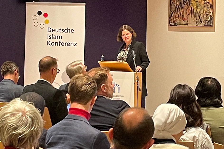 Eine Frau steht hinter einem Rednerpult vor einem Publikum.  (Quelle: BAMF) Eine Frau steht hinter einem Rednerpult vor einem Publikum.