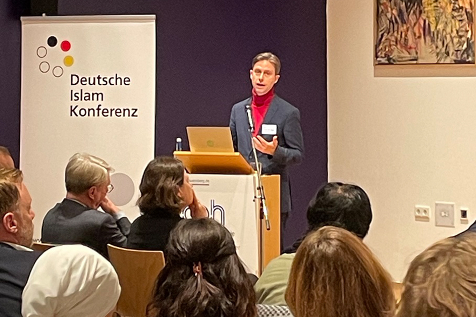 Ein Mann steht hinter einem Rednerpult vor einem Publikum.  (Quelle: BAMF) Ein Mann steht hinter einem Rednerpult vor einem Publikum.