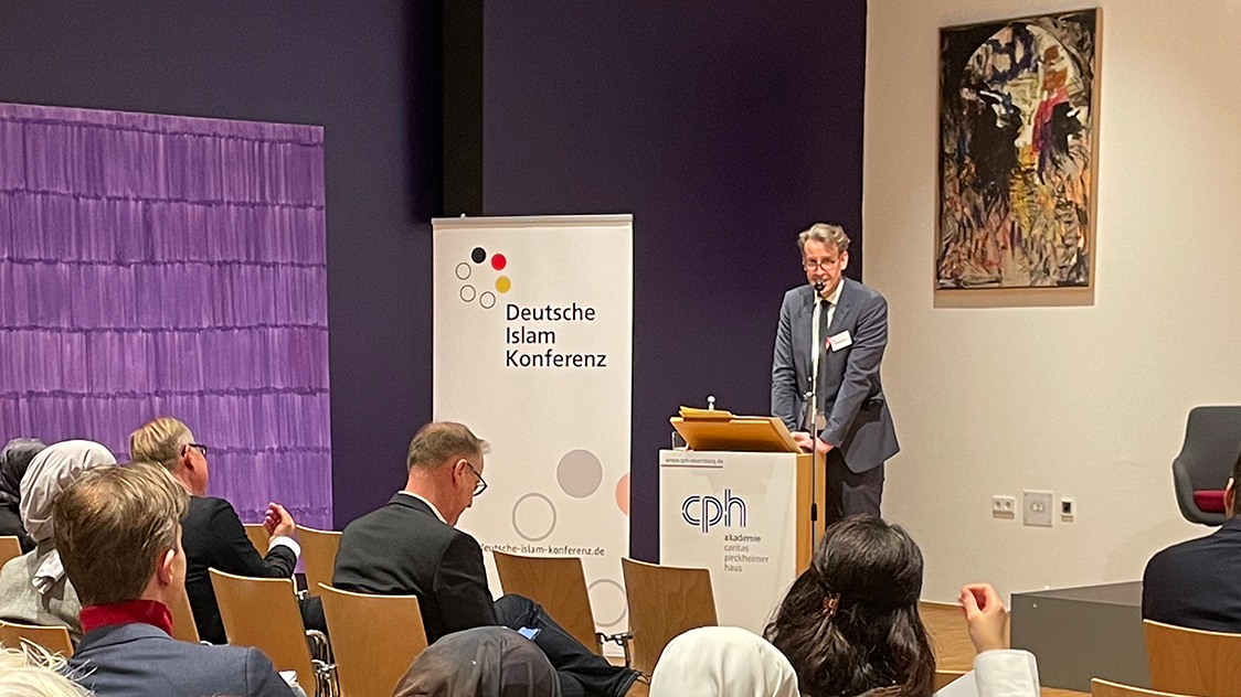 Ein Mann steht hinter einem Rednerpult vor einem Publikum.  (Quelle: BAMF) Ein Mann steht hinter einem Rednerpult vor einem Publikum.