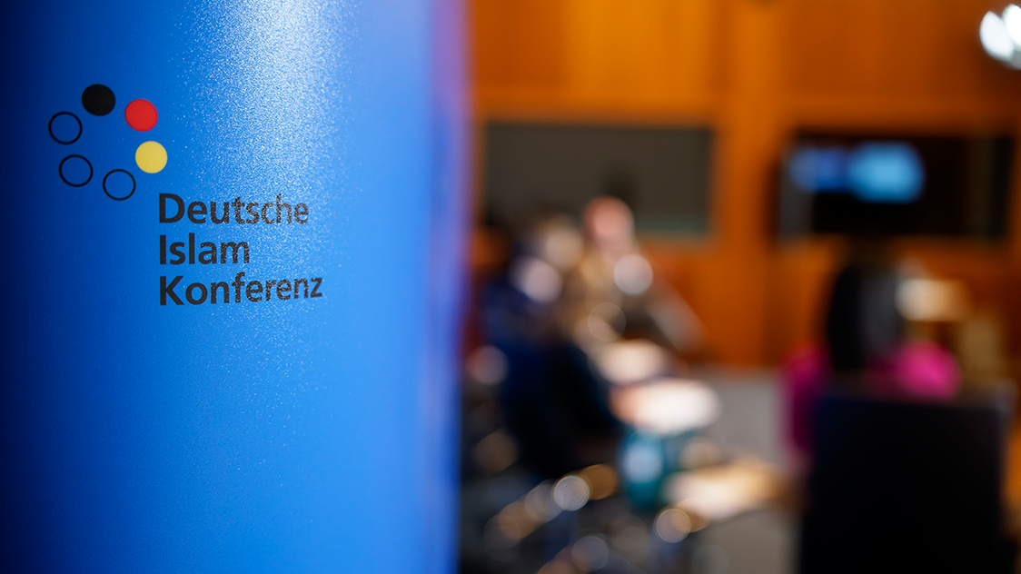 Das Logo der Deutschen Islam Konferenz im Vordergrund und im Hintergrund verschwommen einige Personen, Bildschirme und Stühle.  (Quelle: Henning Schacht) Das Logo der Deutschen Islam Konferenz im Vordergrund und im Hintergrund verschwommen einige Personen, Bildschirme und Stühle.