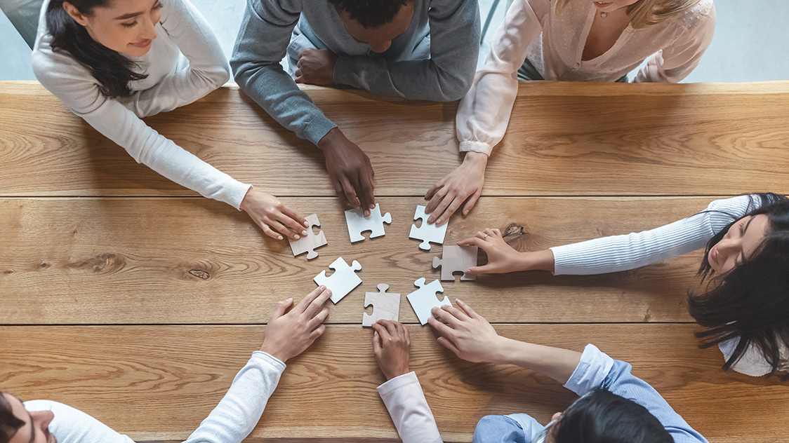 Sieben Personen sitzen an einem Tisch und strecken jeweils den rechten Arm in die Mitte des Tisches (Quelle: AdobeStock | Prostock-studio) Sieben Personen sitzen an einem Tisch und strecken jeweils den rechten Arm in die Mitte des Tisches