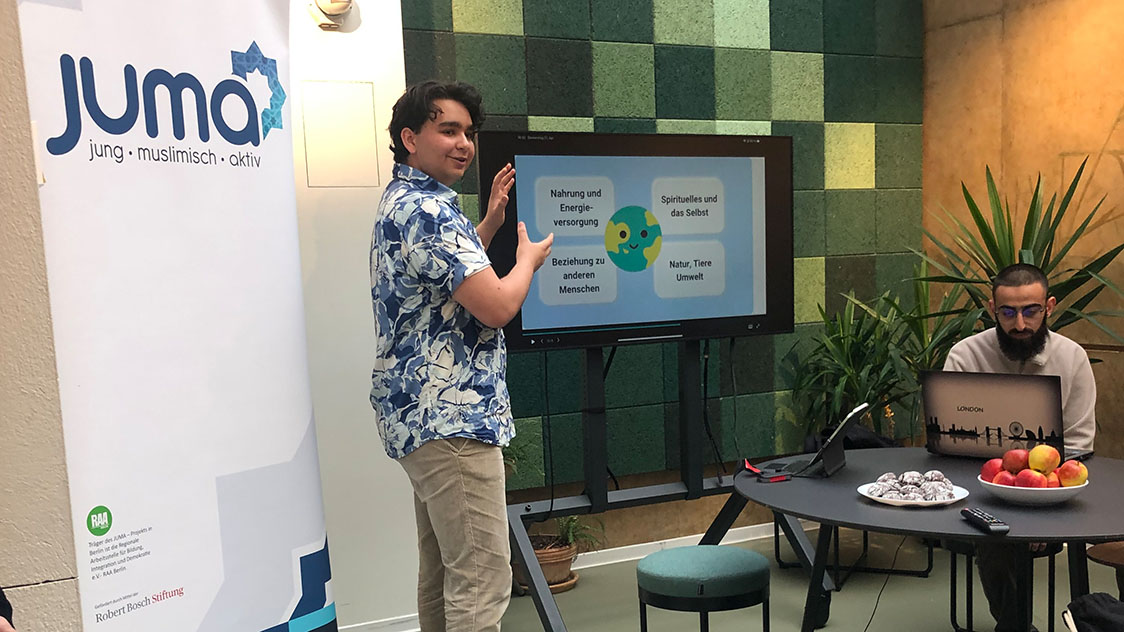 Ein Mann steht vor ein Bildschirm und ein Mann sitzt an einem Tisch (Quelle: JUMA) Ein Mann steht vor ein Bildschirm und ein Mann sitzt an einem Tisch