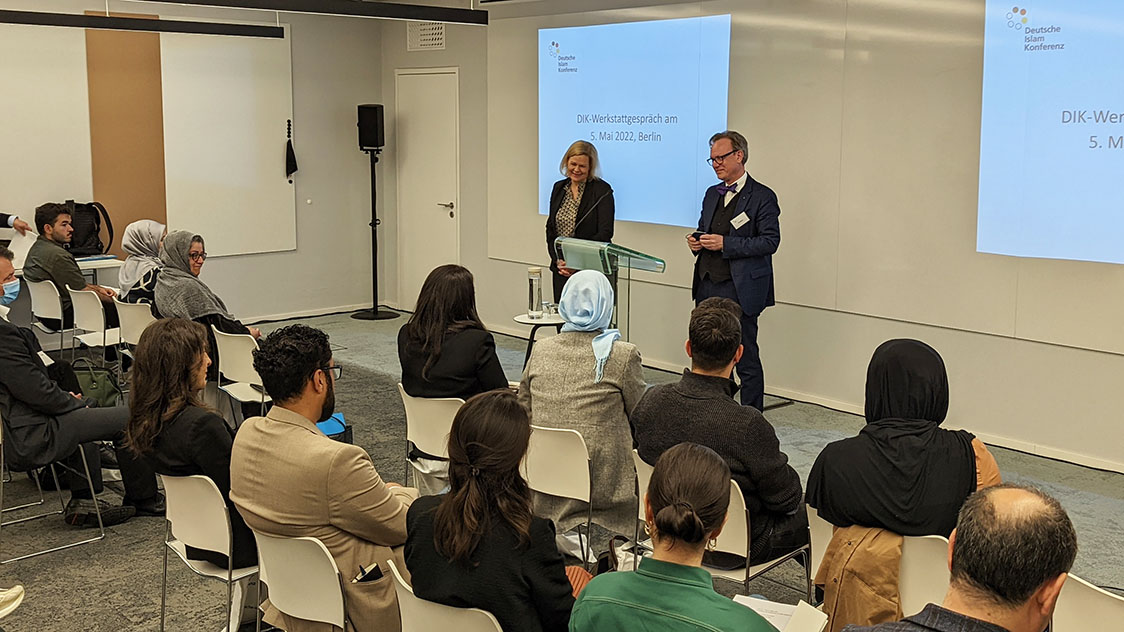 Eine Frau und ein Mann stehen vor einem sitzenden Publikum (Quelle: BAMF) Eine Frau und ein Mann stehen vor einem sitzenden Publikum